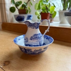 May include: A white and blue ceramic pitcher and bowl set. The pitcher has a curved handle and a detailed blue landscape design. The bowl has a matching design around the rim. The set is placed on a wooden surface.