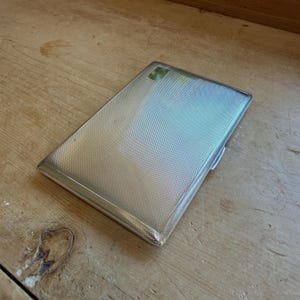 May include: A rectangular silver-toned metal cigarette case featuring a fine, cross-hatched engine-turned texture across its surface. The case has a smooth, polished border and a small, plain rectangular cartouche located in the top corner.