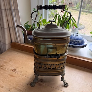 May include: Antique brass kettle with a black wooden handle and a decorative base. The kettle has a spout and a lid with a small knob. The base features ornate cutouts and rests on claw feet. A vintage kitchenware item.