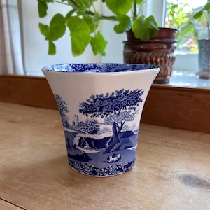 Peut inclure: Un vase en céramique blanc et bleu de forme évasée. Le vase présente une scène de paysage bleue détaillée avec des arbres, des bâtiments et des personnages. Le bord a un motif floral bleu. Le vase est sur une surface en bois.