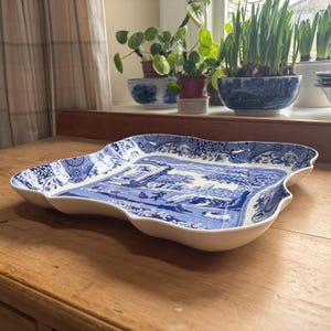 May include: A white and blue ceramic serving tray with a scalloped edge. The tray features a detailed blue landscape design on a white background. The tray is rectangular with a wavy, irregular shape.