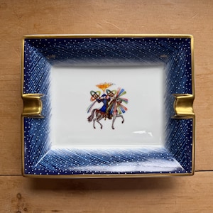 May include: A rectangular porcelain ashtray featuring a deep blue border with white polka dots and diagonal etched lines. The center is white with a colorful illustration of a figure riding a horse, and the edges are trimmed in gold.