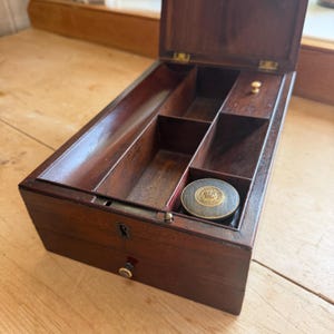 May include: A dark brown wooden writing box with an open lid, featuring multiple interior compartments, a small brass knob, and a circular inkwell with a gold-colored emblem on the lid. The front of the box includes a keyhole and a small drawer.