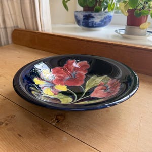 Può includere: Una ciotola rotonda in ceramica con una finitura blu scuro lucida, caratterizzata da un motivo floreale dipinto a mano. L'interno mostra grandi e vivaci fiori di ibisco rossi con accenti gialli e foglie verdi, resi con uno stile pittorico.