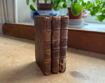 May include: Three antique books bound in brown leather with gold lettering on the spines. The books are stacked vertically on a wooden surface.