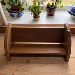 May include: A wooden bookshelf with a warm, brown finish. The shelf has a simple design with a flat base and a back panel. The sides are curved, and there is a small decorative element on one side. The shelf is empty.
