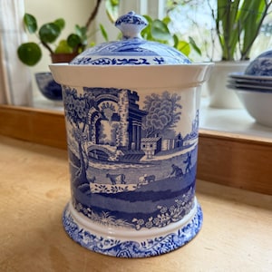 May include: A cylindrical ceramic canister with a matching lid, featuring a blue and white transferware pattern. The design depicts a pastoral landscape with architectural ruins, trees, and figures, finished with a decorative floral border.