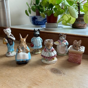 May include: A selection of miniature animal figurines, including a cat, a rabbit with babies, a dog, and a mouse. The figurines are brightly coloured and are displayed on a wooden surface. The figurines are likely made of ceramic or porcelain.