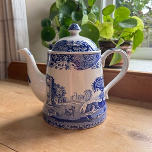 May include: White and blue ceramic teapot with a lid. The teapot features a detailed blue landscape design with figures and buildings. The handle and spout are white. The teapot is sitting on a wooden surface.