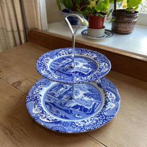 May include: A two-tiered cake stand with blue and white plates. The plates have a scenic design with buildings and trees. The stand has a silver metal handle. The plates are on a wooden surface.