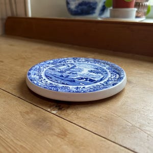 Könnte beinhalten: Ein runder, blau-weißer Keramikuntersetzer mit Landschaftsmotiv. Der Untersetzer hat einen weißen Rand und liegt auf einer Holzoberfläche. Das Design zeigt eine detaillierte blaue Illustration.