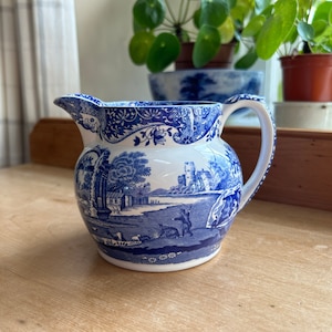May include: A blue and white ceramic jug with a handle. The jug depicts a detailed landscape scene with a castle, figures, and trees. The rim and handle are decorated with floral patterns.