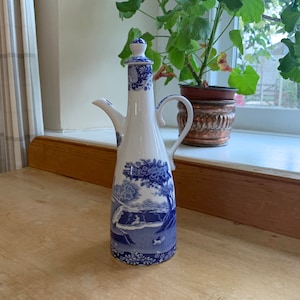 May include: A tall white ceramic cruet or oil dispenser featuring a blue transferware pattern of trees, landscapes, and figures. The vessel has a slender neck, a curved handle, a pointed spout, and a matching removable lid with a small knob.