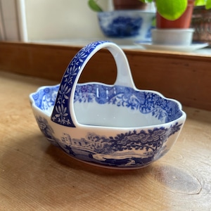 May include: A blue and white porcelain basket with a handle. The basket features a floral design and a scene of a house and trees.