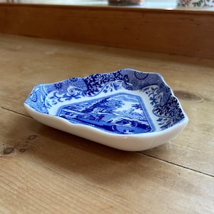 Puede incluir: Un plato triangular de cerámica azul y blanco con borde festoneado. El plato presenta un patrón floral azul y blanco y una imagen central de una escena campestre.