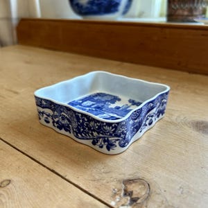 Puede incluir: Plato cuadrado de porcelana con diseño floral y paisajístico azul y blanco. El plato tiene un borde ondulado y un interior blanco. El diseño recuerda a la clásica porcelana azul y blanca.