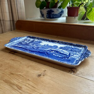 May include: A rectangular ceramic serving tray featuring a blue and white transferware pattern. The design depicts a pastoral landscape with trees, figures, and a building. The tray has decorative handles on each end and a scalloped edge border.