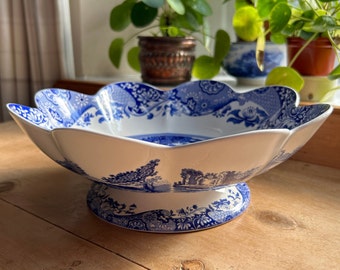 May include: A white ceramic bowl with a scalloped edge and a blue transferware pattern featuring landscape scenes and floral motifs. The bowl is supported by a circular pedestal base decorated with matching blue patterns on a white background.