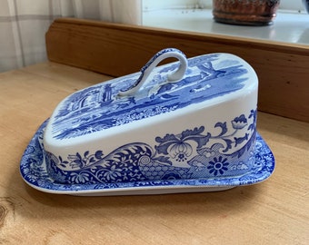 May include: A white and blue ceramic butter dish with a lid. The lid features a blue landscape scene and a curved handle. The base and lid are decorated with floral and scroll patterns. The dish is placed on a wooden surface.