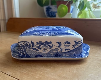 May include: A rectangular ceramic butter dish featuring a classic blue and white transferware pattern. The lid and base are decorated with intricate floral motifs, scrolling vines, and detailed landscape scenes in a deep cobalt blue on white.