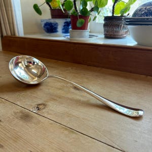 May include: A shiny silver ladle with a rounded bowl and a long, curved handle. The ladle is resting on a wooden surface, reflecting the light. The kitchen utensil is designed for serving soups and sauces.