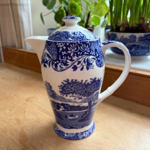 May include: A blue and white ceramic coffee pot with a lid. The pot features a landscape design with trees and a river. The handle and spout are white. The pot is sitting on a wooden surface.