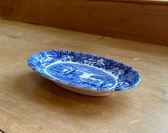 May include: An oval, blue and white ceramic dish with a scalloped edge. The dish features a landscape scene in the center and decorative patterns around the rim. The dish is resting on a wooden surface.
