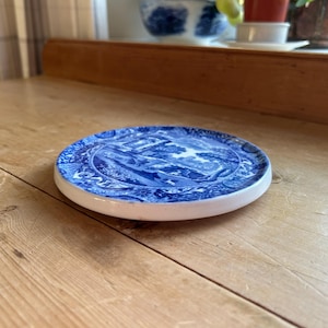 Peut inclure: Un dessous de verre rond en céramique bleue et blanche avec un motif de paysage. Le dessous de verre a un bord blanc et est posé sur une surface en bois. Le motif bleu représente des arbres, des bâtiments et de l'eau. Un objet décoratif pour la maison.