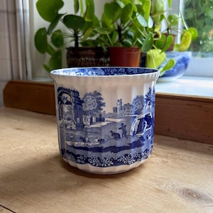 May include: A white ceramic planter with vertical ribbing and a blue transferware pattern depicting a pastoral landscape with figures, trees, and architectural ruins. The interior rim features a decorative blue floral border design.