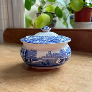 Op de afbeelding: Een witte suikerpot van keramiek met een blauw en wit bloemenmotief. De pot heeft een deksel en is versierd met een scène met een kasteel en bomen.