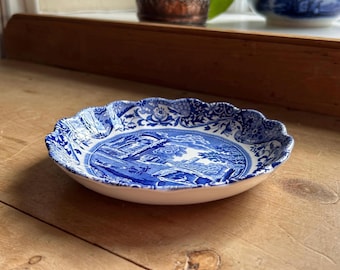 May include: A small, round, white and blue decorative plate with a scalloped edge. The plate features a blue landscape scene in the center and floral patterns around the rim. The plate is made of ceramic.