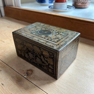 Könnte beinhalten: Eine rechteckige antike Box mit einem floralen Design in Gold und Schwarz. Die Box hat einen Klappdeckel und steht auf einer Holzoberfläche. Die Oberseite der Box hat ein kreisförmiges Design mit einem kursiven Buchstaben.