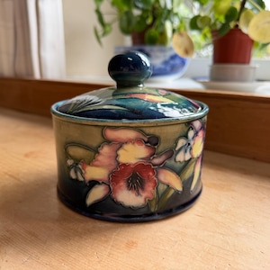May include: A round ceramic lidded jar featuring a dark blue and sage green glaze. The exterior is decorated with a hand-painted floral motif in shades of pink, yellow, and white. The lid has a matching dark blue knob handle on top.