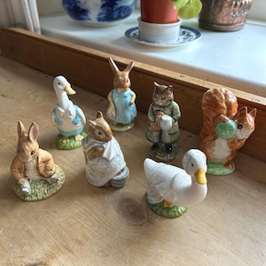 May include: A selection of ceramic animal figurines, including a rabbit, a duck, a squirrel, and a cat. The figurines are painted in detail and are set on a wooden surface. The figurines are in various poses and outfits.