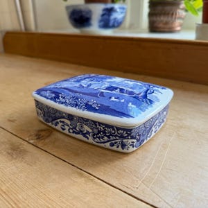 Puede incluir: Una caja rectangular de cerámica con base blanca y patrones azules detallados. La tapa muestra una escena de paisaje con figuras y follaje, mientras que los lados están decorados con un motivo floral que se repite en toda la pieza.