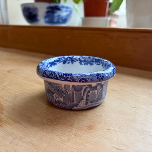 May include: A small, round, blue and white ceramic bowl with a detailed landscape pattern. The bowl has a wide, rounded rim and a white interior. The design features buildings, trees, and other elements in a classic, vintage style.