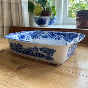 Puede incluir: Una fuente rectangular de cerámica blanca con diseños florales y paisajísticos azules. La fuente tiene un borde festoneado y está sobre una superficie de madera. El diseño incluye árboles, edificios y figuras.