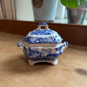 Puede incluir: Un cuenco con tapa de cerámica azul y blanca con un diseño de paisaje. El cuenco tiene forma hexagonal, asas pequeñas y descansa sobre tres patas. La tapa presenta un asa de bucle. El diseño es de estilo transfer clásico azul y blanco.