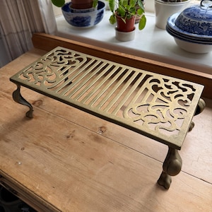 Op de afbeelding: Een rechthoekige messing trivet met sierlijke, opengewerkte ontwerpen en gebogen poten. De trivet heeft een reeks parallelle staven over de bovenkant en decoratief krulwerk aan de uiteinden. Het is ongeveer 25 cm lang.