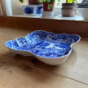 May include: A white and blue ceramic serving dish with a scalloped edge. The dish features a blue and white pastoral scene. The dish is on a wooden surface.