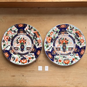 May include: Two matching decorative plates with a floral design. The plates feature a navy blue border with orange and white floral patterns. The center of each plate has a vase with flowers. The plates are on a wooden surface.
