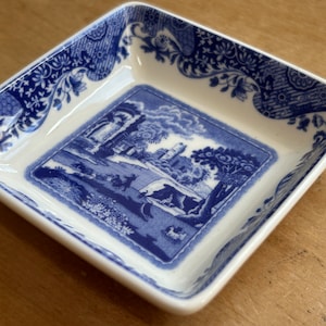 May include: A small, square, white ceramic dish with a blue and white floral pattern. The center of the dish features a blue and white scene of a building and trees.