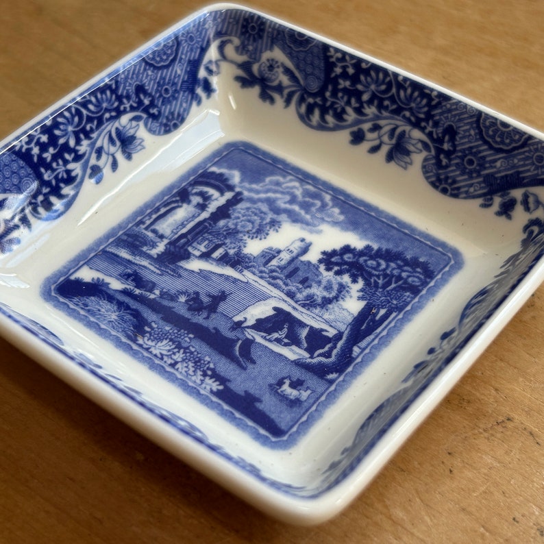 May include: A small, square, white ceramic dish with a blue and white floral pattern. The center of the dish features a blue and white scene of a building and trees.