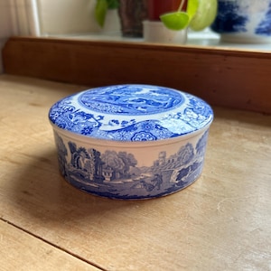 Könnte beinhalten: Eine runde, blau-weiße Keramik-Schmuckdose mit Deckel. Die Dose und der Deckel weisen ein detailliertes Landschaftsdesign mit Bäumen, Gebäuden und Figuren auf. Die Dose steht auf einer Holzoberfläche.