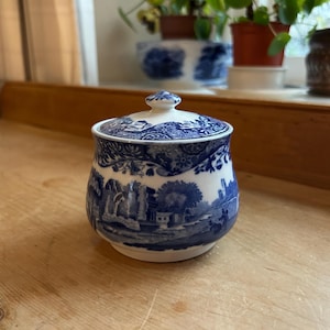 Puede incluir: Un azucarero de cerámica azul y blanco con tapa. El cuenco presenta un diseño de paisaje con árboles y edificios. La tapa tiene un patrón floral y un pequeño pomo. El azucarero está sobre una superficie de madera.