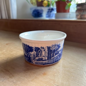 Puede incluir: Un cuenco pequeño de cerámica blanca con un diseño de paisaje azul en el exterior. El cuenco tiene un borde ligeramente ensanchado y un acabado liso y brillante. El diseño azul representa árboles, edificios y elementos acuáticos.