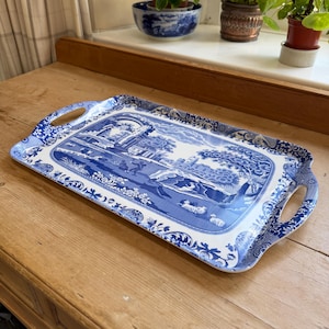 Könnte beinhalten: Ein rechteckiges Serviertablett mit einem blau-weißen Transferdruckmuster. Die zentrale Szene zeigt eine ländliche Landschaft mit Figuren, Bäumen und Gebäuden, umrahmt von einer floralen Bordüre und zwei integrierten seitlichen Griffen.