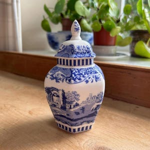 May include: A white and blue ceramic jar with a lid. The jar features a detailed landscape scene and floral patterns. The lid has a decorative finial. The jar is a decorative item.