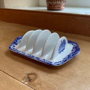 Peut inclure: Un porte-toast en céramique blanche avec quatre fentes, orné d'un motif floral bleu et blanc. Le porte-toast est conçu pour contenir quatre tranches de pain grillé.