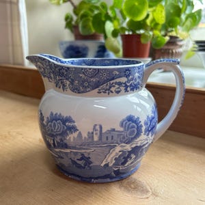 Puede incluir: Jarra de cerámica blanca y azul con diseño de paisaje. La jarra presenta un detallado patrón azul alrededor del borde y el asa, con una ilustración escénica en el cuerpo. El asa es curva y el pico es puntiagudo.